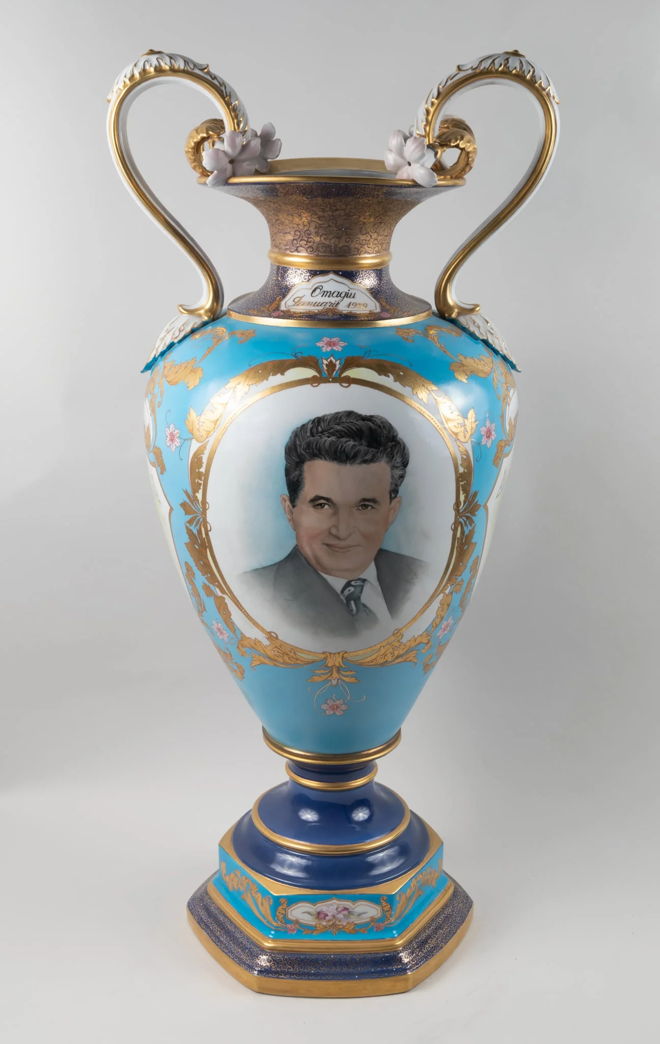 Vază din porțelan, fond albastru, tip amforă, cu anse, decorată cu două medalioane, unul reprezentând portret Nicolae Ceaușescu, cel de-al doilea un buchet cu flori. Pe gâtul vazei o dedicație ”Omagiu, Ianuarie 1989”. Vază oferită lui Nicolae Ceaușesacu de Consiliul Național al Femeilor cu ocazia aniversării zilei de naștere, ianuarie 1989., detail