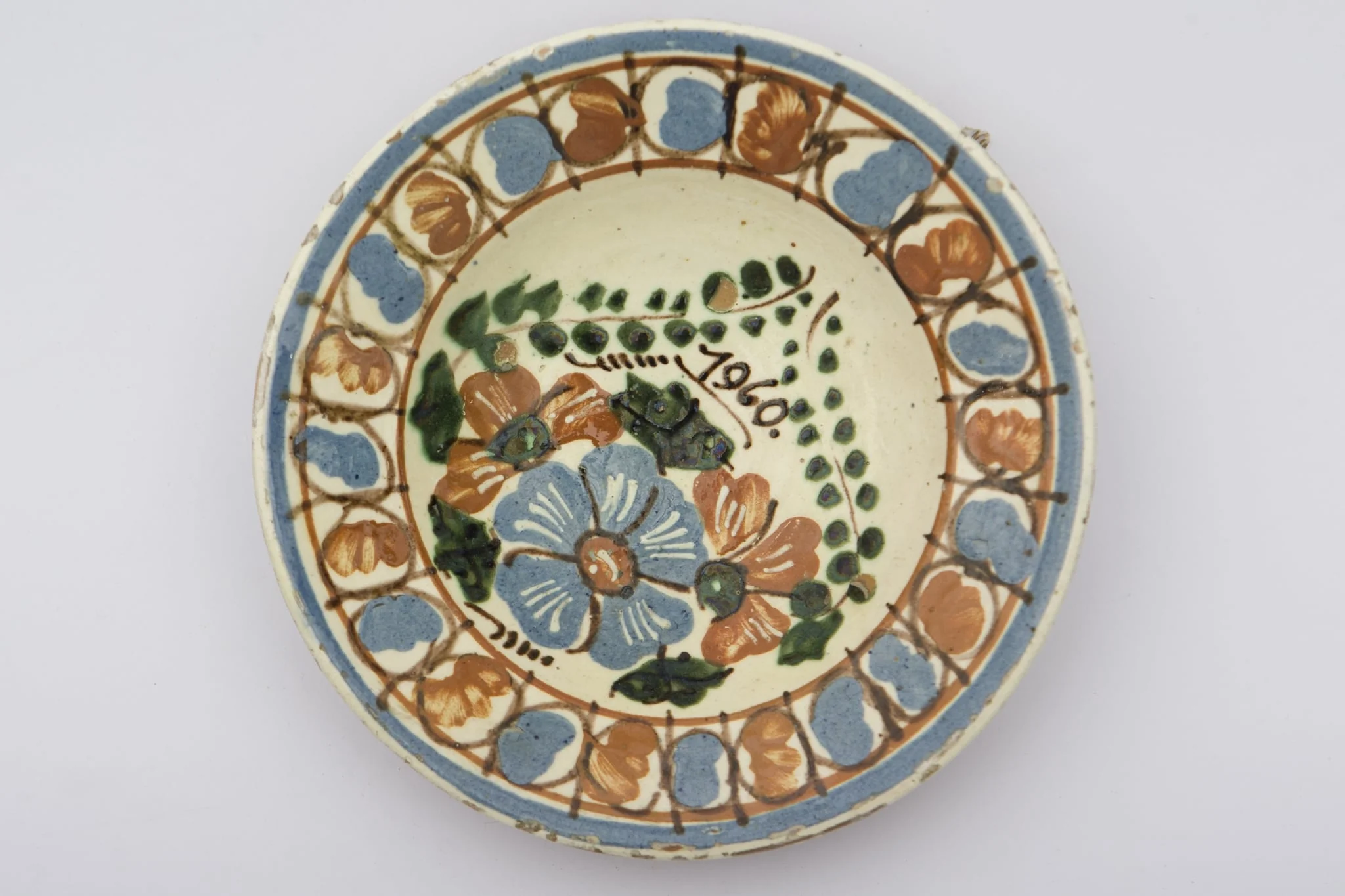 Recipient ceramic smălțuit, de formă tronconică, cu baza plată, pereții ușor ridicați și buza profilată. Peste stratul alb de angobă au fost realizate două registre ornamentate cu motive geometrizante și fitomorfe stilizate. Primul registru, situat pe fundul vasului este format din trei flori: una, centrală cu mijlocul maro și petalele albastre, celelalte două de culoare maro roșcat. Crenguțelor, pe care sunt poziționate florile, li se unesc vârfurile, deasupra cărora a fost trecută datare: „1960”. Marginal a fost realizat cel de-al doilea registru, format din boboci de culoare albastru și maro roșcat, despărțiți de linii verticale drepte. Două linii circulare drepte încadrează ultimul registru. Farfuria era utilizată în gospodărie pentru decorarea interioarelor. Prezintă uzură funcțională. Cromatică: fond: alb; motive: albastru; maro roșcat; verde., detail