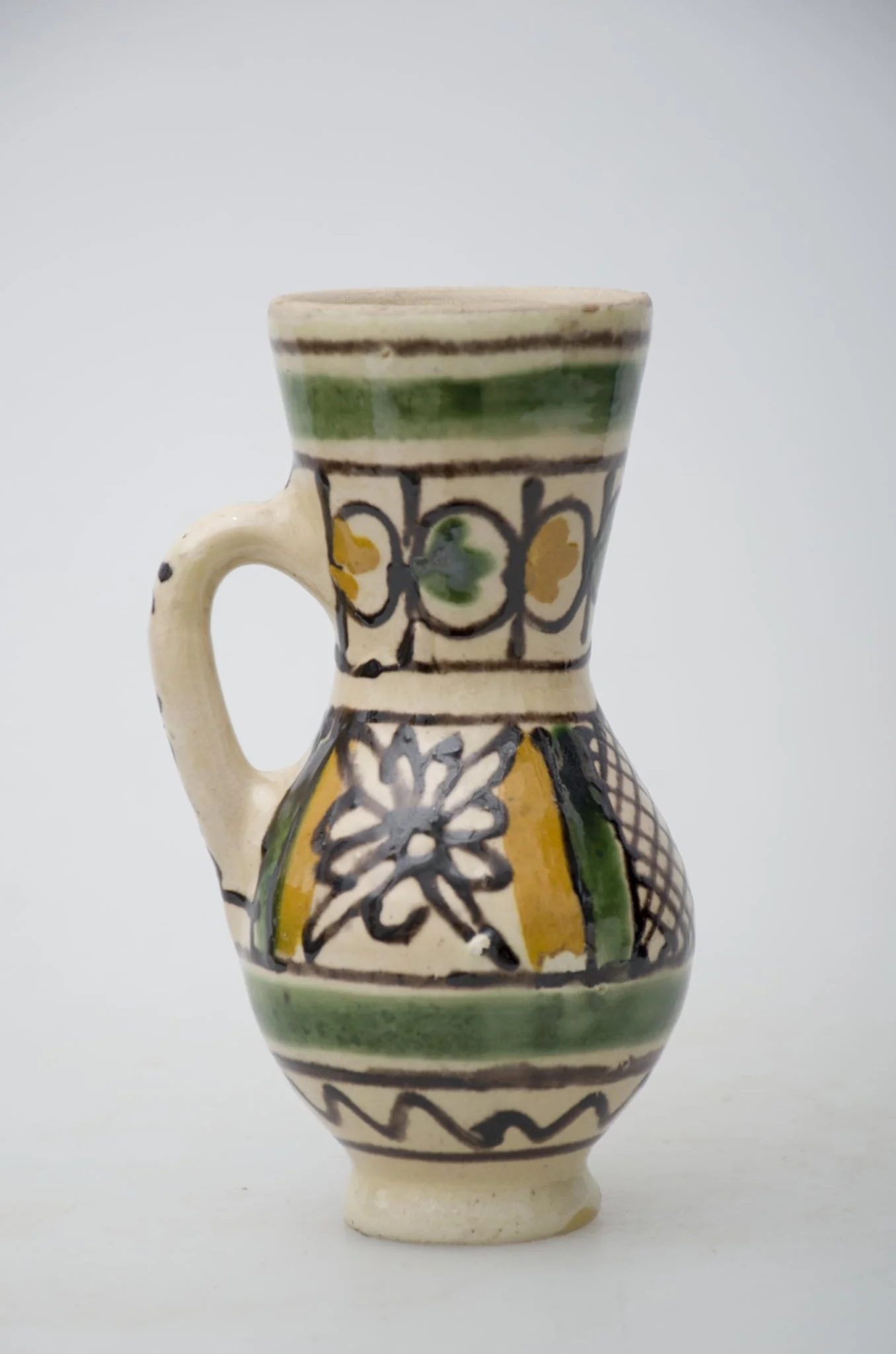 Vas ceramic de formă bitronconică, smălțuit de mici dimensiuni, cu baza plată și bombat în zona „pântecelui”. Prezintă o toartă ce pornește din zona gâtului redus ca dimensiuni și se termină în zona diametrul maxim al vasului. Decorul vasului este compus din două registre picturale. În primul registru, mărginit de două linii drepte subțiri, de culaore maro închis, au fost pictate două flori conturate cu negru și încadrate de câte două linii groase de culoare verde și galben. Cel de-al doilea registru este compus din boboci de culoare verde și galben, fiind dispuși concentric pe gâtul vasului. Ambele registre picturale sunt încadrate între două linii groase circulare, verzi. Pe toartă a fost redactat textul: „KORUND”. Prezintă uzură funcțională. Cromatică: fond: alb; motive: galben, verde, maro închis., detail