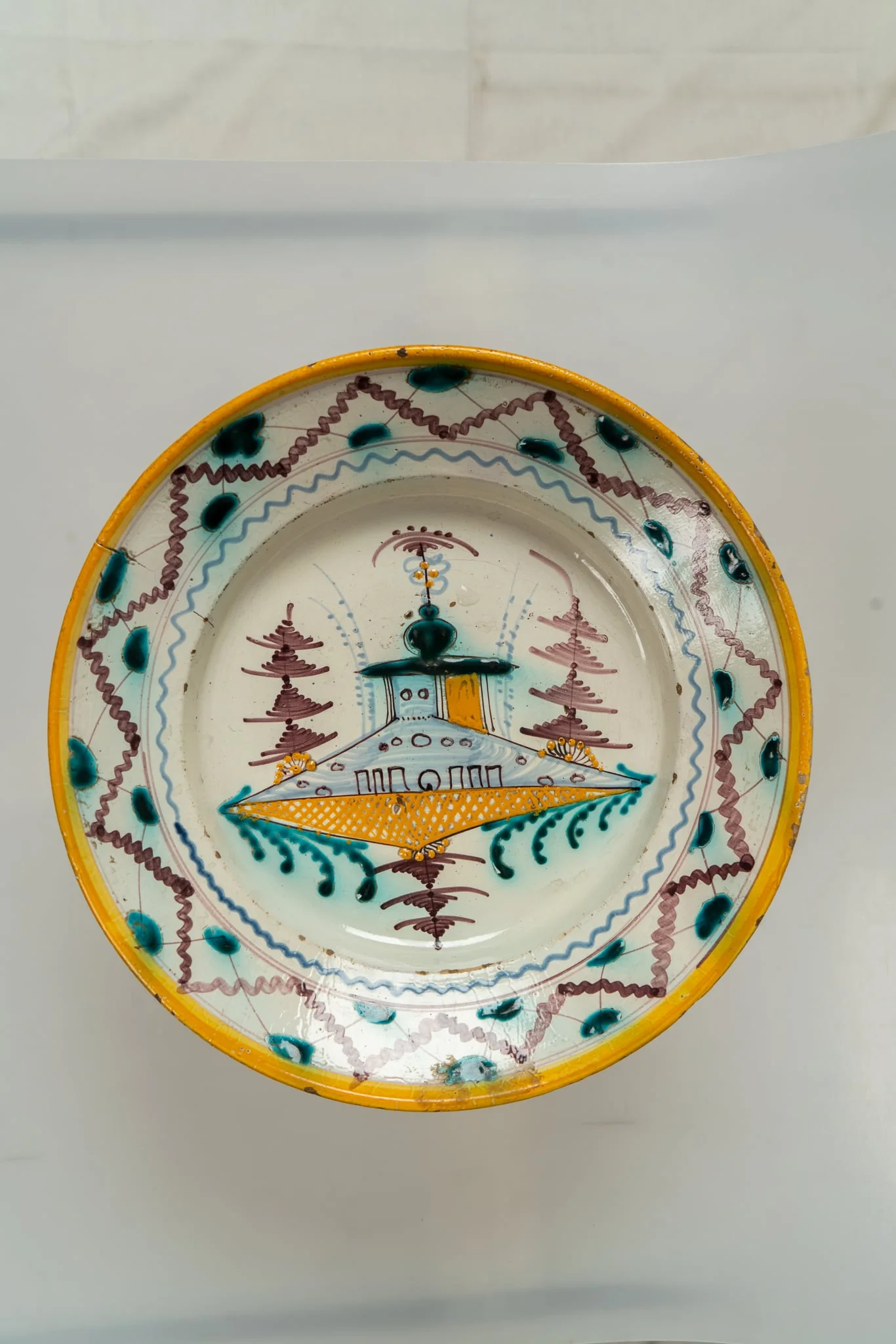 Farfurie de formă aplatizată, cu marginea lată, răsfrântă. Decorul principal este dispus în centrul farfuriei, pe un fond alb-lăptos, și prezintă un motiv arhitectural și vegetal stilizat, iar buza farfuriei este decorată cu motive geometrice (două linii vălurite dispuse în zig-zag, alternativ, una mai groasă, cealaltă mai subțire, de culoare maro; pete de culoare verde turcoaz, linii subțiri dispuse sub formă de cercuri concentrice). Muchia buzei este marchată de o dungă galbenă. Cromatica: alb, maro, galben ocru, albastru deschis, verde turcoaz., detail