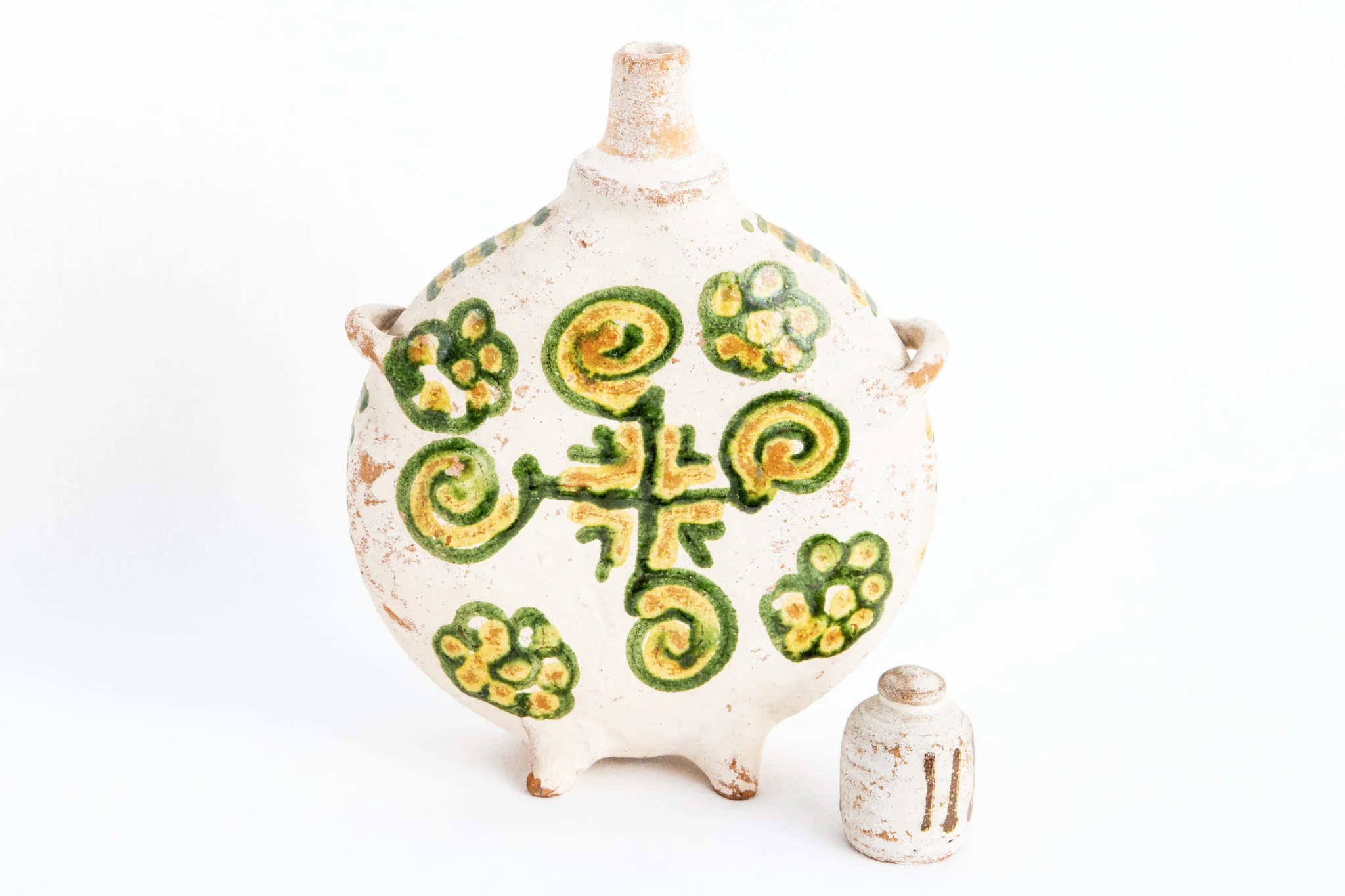 Ploscă de dimensiuni mici prevăzută cu capac. Fond alb. Decorul este similar pe ambele fețe ale obiectului; o cruce de culoare verde dispusă central; între brațele crucii sînt amplasate unghiuri duble colorate în galben respectiv, verde. Crucea este încadrată de patru spirale colorate în galben și verde și patru flori stilizate realizate cu aceleași culori. Muchia ploștii este decorată cu linii orizontale de culoare galbenă și verde, în alternanță cromatică., detail