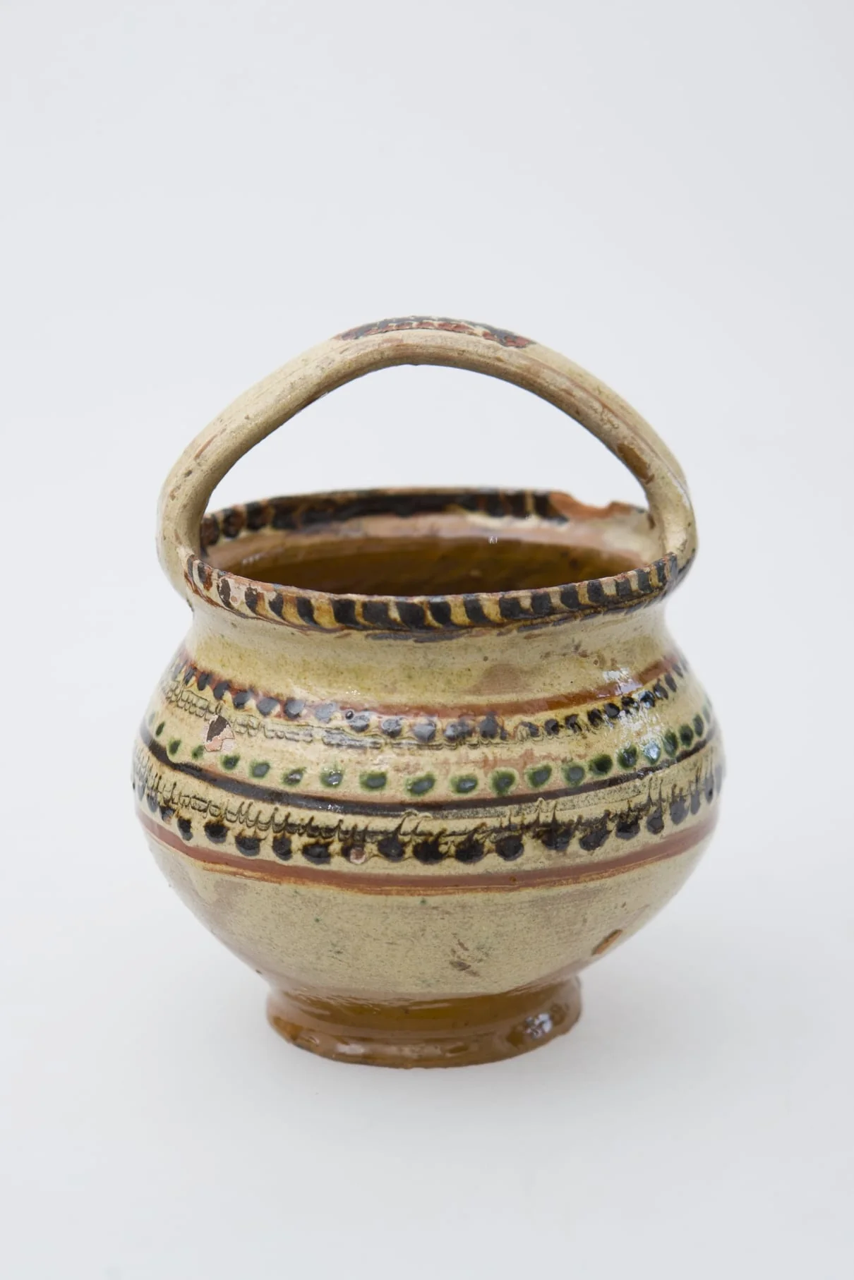 Vas ceramic de mici dimensiuni, pe bază inelară, pântecos, cu fundul și gura late. Deasupra gurii vasului a fost pusă o toartă de prindere. Buza vasului a fost pictată printr-un brâu de crenguțe. Pe pântecul vasului brâuri de puncte și două benzi subțiri jirăvite. Smălțuit în interior și exterior. Cromatică: fond: crem; motive: verde; maro; bej., detail