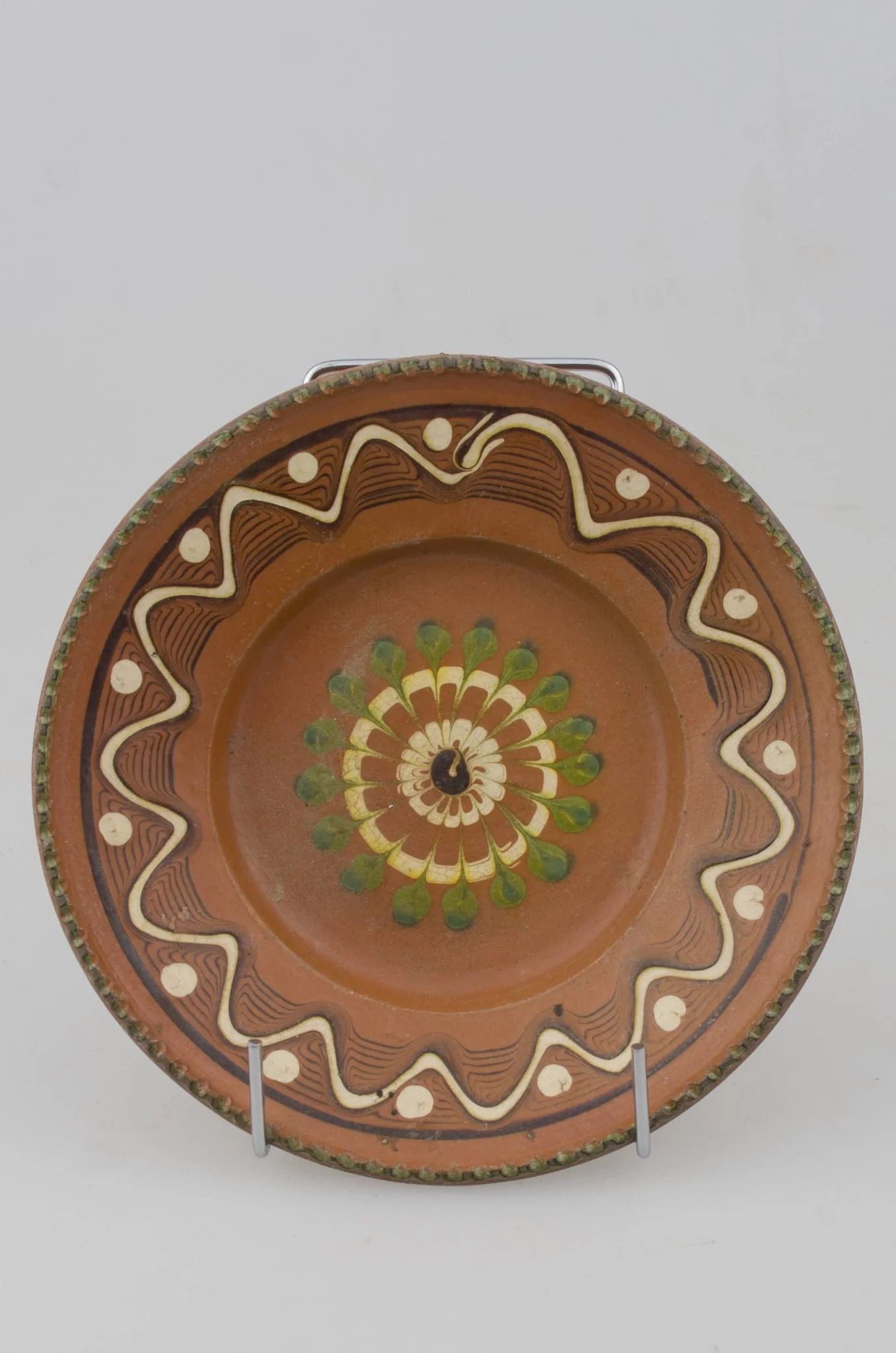Farfurie cu pereții rotunjiți, marginea oblică lată și buza crestată. Prezintă decor geometric pe margine: linii frânte și curbe; pete de culoare; central avem o floare stilizată. Cromatică: fond: maro; motive: verde; alb., detail