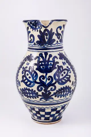 Cană piriformă prevăzută cu o toartă dreaptă și lată, gura rotundă, prevăzută cu cioc de scurgere. Suprafața decorativă a vasului este împărțită în patru registre decorative. Primul registru dispus pe gâtul piesei prezintă un decor floral și fitomorf reprezentând lalele și lujeri. Al doilea registru prezintă o linie vălurită și trei linii subțiri circulare. Decorul central, reprezintă trei „arbori ai vieții” stilizați realizați din trei lalele mari, înflorite, lujeri și frunze, separați de câte o semilună. Registrul al patrulea prezintă motivul tablei de șah. Deasupra toarte pictate cu motive florale și fitomorfe este redat anul confecționării piesei: 1980, iar pe fundul vasului numele persoanei care a pictat piesa: Herta Wilk. Cromatica: alb, albastru-cobalt.