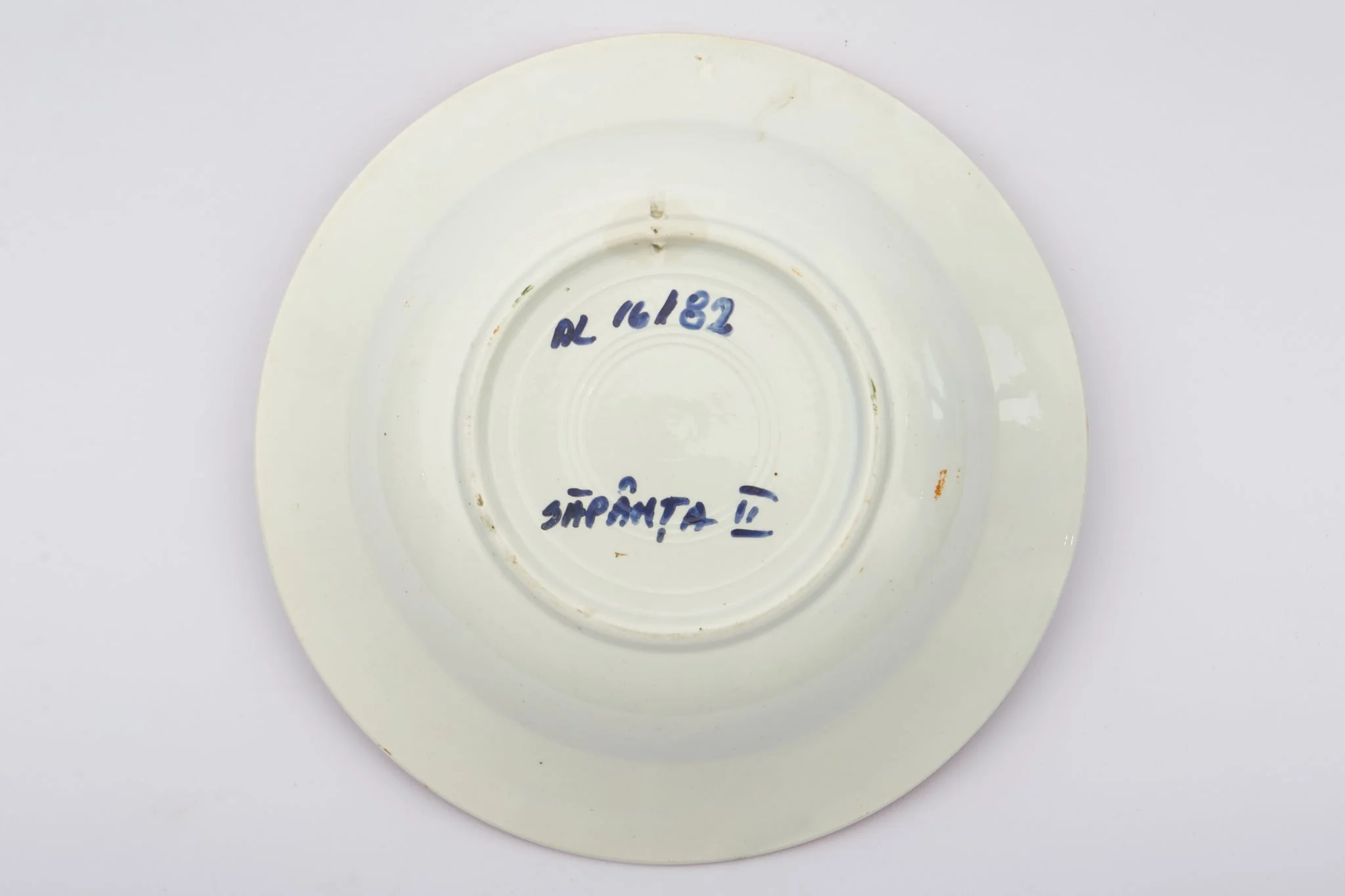Vas ceramic de dimensiuni reduse, având formă tronconică, cu baza plată, pereții reficați și marginile evazate. Peste fondul alb au fost realizate decorațiile geometrizante și fitomorfe stilizate, dispuse în două registre. Primul registru este format dintr-un trandafir portocaliu cu frunze verzi, având în laterale câte o floare cu mijlocul albastru și petalele roșii, și câte un boboc roșu. Al doilea registru este format dintr-o tulpină circulară verde, pe care se dezvoltă trei grupuri de flori mici albastre. Alte trei grupuri, a câte trei linii portocalii oblice, însoțesc decorul fitomorf. Acest ultim registru este încadra dinspre interior de bandă groasă portocalie. Buza farfuriei a fost la rândul ei conturată cu portocaliu. Prezintă uzură funcțională. Cromatică: fond: alb; motive; portocaliu; verde; albastru., detail