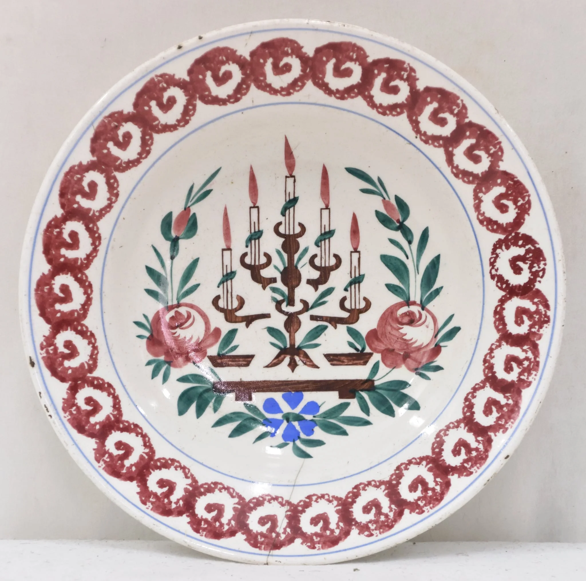 Obiectul are o formă tronconică cu baza mică în partea inferioară. Decorul se află numai la interiorul farfuriei fiind pictat cu motive geometrice, florale, vegetale și un simbol religios: pe fundul farfuriei a fost pictată o menoră cu cinci brațe (sfeșnic - obiect de cult evreiesc) sprijinită pe un suport; fiecare dintre brațele sfeșnicului susține câte o lumânare aprinsă; această reprezentare este încadrată de tulpini, frunze, flori și boboci (trei flori plus doi boboci); întreaga pictură de pe fundul farfuriei este încadrată de un cerc iar un altul este "trasat" de jur-împrejur în partea superioară, la buza piesei; între cele două cercuri se găsește un șirag format din cercuri (cu diametre egale) tangente la exterior, asemănător unui lanț cu zale rotunde; fiecare dintre aceste cercuri are la interior un semn, ce amintește de litera "V". Pe spatele farfuriei se află un mic orificiu rotund prin care se introduce firul textil pentru posibilitatea atârnării obiectului de perete. Culoarea piesei este albă iar pictura este realizată cu următoarele culori: maro închis, maro deschis, bleu și verde., detail