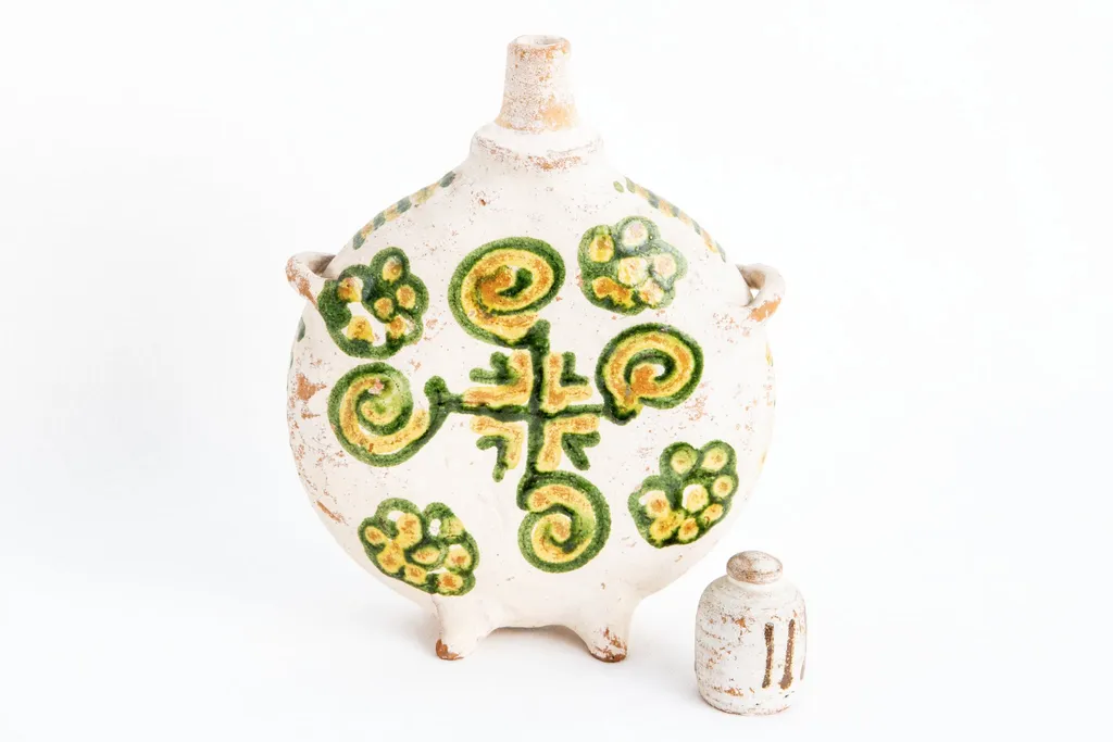 Ploscă de dimensiuni mici prevăzută cu capac. Fond alb. Decorul este similar pe ambele fețe ale obiectului; o cruce de culoare verde dispusă central; între brațele crucii sînt amplasate unghiuri duble colorate în galben respectiv, verde. Crucea este încadrată de patru spirale colorate în galben și verde și patru flori stilizate realizate cu aceleași culori. Muchia ploștii este decorată cu linii orizontale de culoare galbenă și verde, în alternanță cromatică., by Dumitru Șchiopu