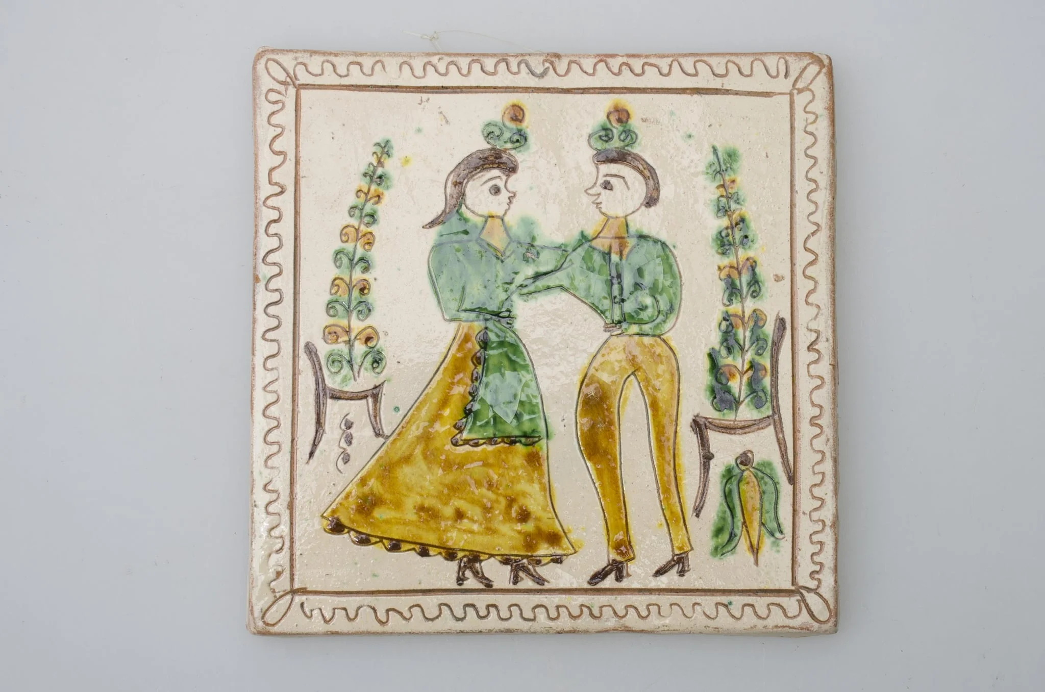 Piesă ceramică de formă pătrată, smălțuită și angobată, cu rol decorativ. Laturile pătratului sunt marcate de două chenare, primul vălurit, al doilea în linie dreaptă. Ornamentată cu motive antropomorfe și floral-vegetale: doi tineri, un băiat și o fată ținându-se pe după mijloc (băiatul cu mâna dreaptă, fata cu stânga). În spatele lor câte un scaun din care se înalță o floare. Motivele au fost la început incizate, iar apoi pictate. Pe spate, cahla este prevăzută cu un orificiu pentru prinderea aței. Tot aici, este inscripționat numele meșterului olar, "Colibaba". Cromatică: fundal alb; motive: verde, galben, cărămiziu, maro, negru., detail