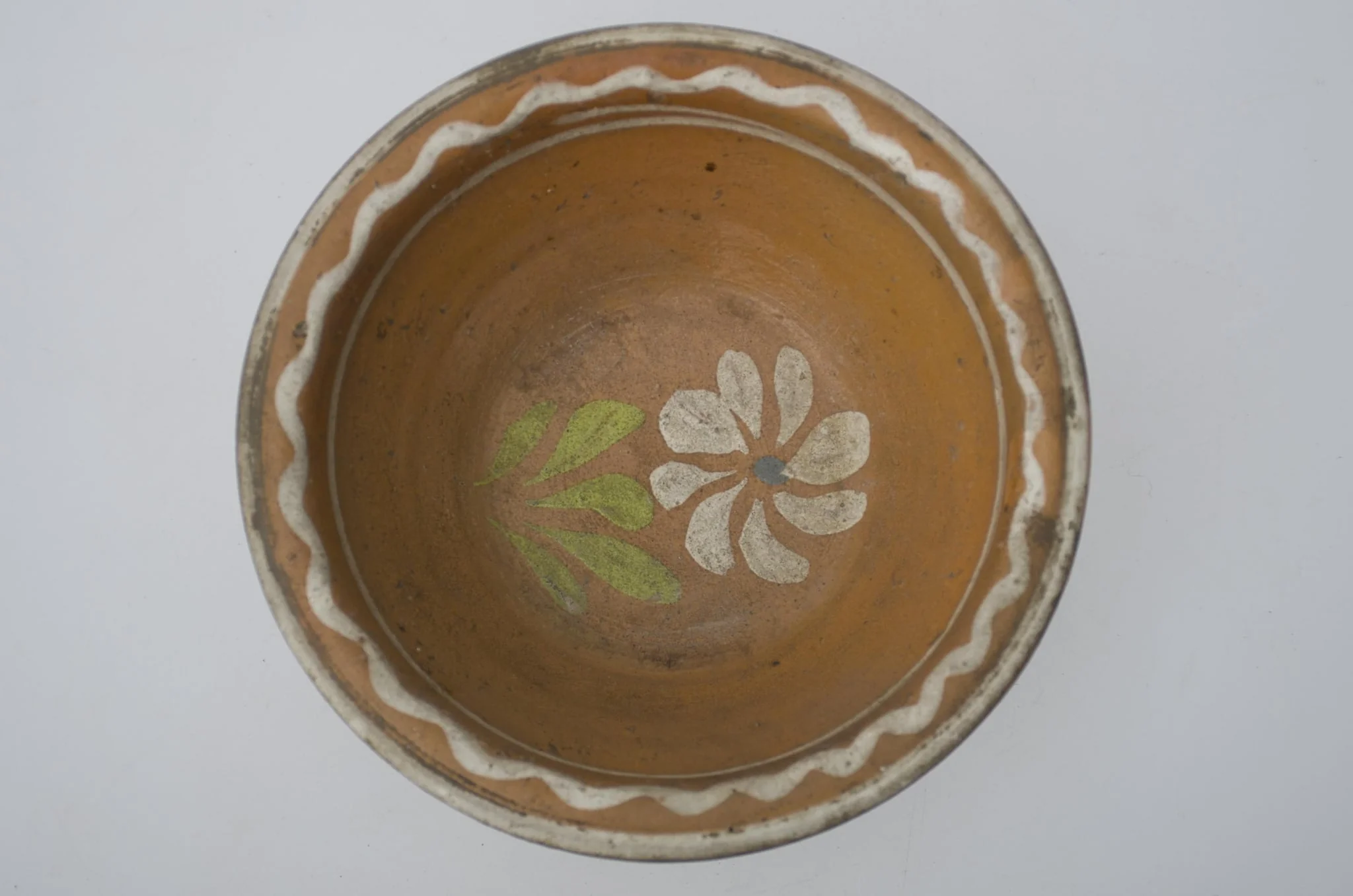 Farfurie adânca, de formă tronconică, smălțuită în interior. Pe margine prezintă decor geometric: linia vălurită; central o floare. Cromatică: fond: cărămiziu; motive: alb; verde; albastru., detail