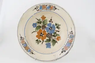 Farfuria este lucrată la roată, prezintă marginea evazată în plan orizontal fiind smălțuit în interior. Pe suprafața interioară a vasului se identifică registre decorative floral-vegetale dispuse pe fundul vasului și pe marginea interioară. Registrele sunt delimitate de două cercuri realizate prin pictare și dispuse pe buza vasului și spre interiorul acestuia. Cromatică: fond: alb; motive: roșu; albastru; verde; brun; maro., 1850