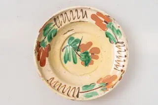 Farfurie adâncă, angobată pe fața interioară cu un strat de humă albă. Este decorată prin pictare cu pensula și cu cornul cu motive florale, fitomorfe și geometrice: central pe fundul farfuriei este reprezentată o floare stilizată, cu petale de culoare siena arsă și frunze verzi, iar buza farfuriei este decorată cu trei flori de culoare maro-roșcat, siena arsă și galben ocru, cu frunze verzi; între flori sunt pictate trei motive formate din câte o linie șerpuită de culoare maro. Cromatica: alb, maro-roșcat, maro, verde, siena arsă, galben ocru. Suprafața ornamentată a farfuriei este acoperită cu un strat de smalț transparent., 1850