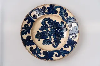 Farfurie adâncă, cu buza evazată; este angobată pe fața interioară cu un strat de humă albă, peste care este pictat cu pensula, cu linii groase, decorul monocromatic format din motive florale și fitomorfe stilizate (tulpină cu o floare de lalea și frunze, reprezentate pe centrul farfuriei și un șir continuu compus din elemente vegetale, ce decorează buza acesteia). Cromatica: alb, albastru. Farfuria este acoperită pe fața decorată cu un strat de smalț transparent., 1850