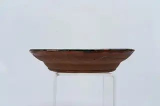 Farfurie adâncă, cu buză evazată; este angobată pe fața interioară cu un strat de angobă maro, peste care este pictat cu cornul, pensula și cu gaița decorul format din motive geometrice: central pe fundul farfuriei este pictată o linie spiralată rotundă de culoare albă; bordura interioară a farfuriei este decorată cu linii circulare concentrice de culoare albă trase cu gaița și cu puncte verzi, iar buza farfuriei cu o linie ondulată circulară și puncte groase de culoare albă. Muchia buzei este marcată cu o dungă verde. Suprafața ornamentată a farfuriei este acoperită cu un strat de smalț transparent. Cromatica: maro, alb, verde., 1850