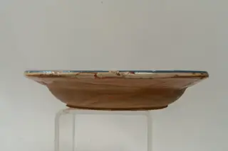 Farfurie adâncă, cu buză răsfrântă, modelată la roata olarului și angobată cu humă albă pe fața interioară a acesteia. Decorul, realizat prin pictare cu pensula și cu cornul, se compune din motive avimorfe (cocoș) și florale stilizate, dispuse într-un registru decorativ în zona centrală a farfuriei. Decorul este completat de câteva motive geometrice (linie ondulată, puncte, linii hașurate, linii sub formă de cercuri concentrice). Pe buza farfuriei: două cercuri concentrice, subțiri și o dungă albastră pe toată lungimea muchiei buzei. Cromatica: alb, albastru, verde, maro, siena naturală, roz pal. Peste decor, pe fața interioară a farfuriei este așezat un strat de smalț transparent., 1850