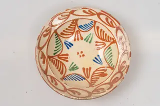 Farfurie adâncă, cu buză relativ îngustă, evazată; este acoperită pe suprafața interioară cu un strat de angobă albă, peste care este pictat cu cornul decorul format din motive geometrice: fundul farfuriei și peretele acesteia este decorat cu motive formate din linii curbe de culoare maro, ce au pe suprafața interioară linii oblice paralele, inegale ca mărime, de culoare verde, albastru, maro; grupuri de câte trei linii colorate, în formă de V și patru puncte centrale, de culoare maro. Buza farfuriei este decorată cu un șir continuu din motive formate din linii ovale, de culoare maro, cu câte două linii sub formă de virgulă în interior. Suprafața ornamentată a farfuriei este acoperită cu un strat de smalț transparent. Cromatica: alb, albastru, verde, maro.