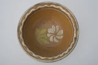 Farfurie adânca, de formă tronconică, smălțuită în interior. Pe margine prezintă decor geometric: linia vălurită; central o floare. Cromatică: fond: cărămiziu; motive: alb; verde; albastru.