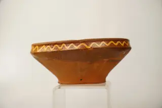 Farfurie adâncă de tip castron, cu fund îngust, corp tronconic și buză verticală, îngustă și ușor profilată. Decorul este pictat cu cornul, cu humă albă, direct pe suprafața neangobată a vasului și se compune din motive geometrice și floral-geometrizate: centrul farfuriei este decorat cu două cercuri concentrice de culoare albă, ce conțin între ele o linie circulară ondulată, iar în mijloc un element decorativ neregulat, de formă palmată, cu puncte albe în interior. Cinci motive aproximativ identice, formate din linii ovale cu puncte în interior și linii mici arcuite pe exterior, sunt pictate pe peretele vasului, pe exteriorul cercului ce marchează fundul acestuia, sugerând niște petale de floare. Patru pete de culoare verde-închis realizate completează decorul din această zonă. Buza farfuriei este decorată pe interior și pe exterior cu o linie circulară ondulată, de culoare albă. Suprafața ornamentată a farfuriei este acoperită cu un strat de smalț transparent. Cromatica: alb, verde-închis, pe fondul roșu al lutului ars., 1850