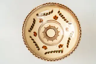 Farfurie adâncă, de tip castron, modelată la roata olarului, angobată pe fața interioară cu humă albă. Decorul este realizat prin pictare cu cornul și prin jirăvire: culorile sunt picurate cu cornul și trase (jirăvite) cu gaița. Motivele decorative sunt florale și vegetale stilizate și decor specific tehnicii prin jirăvire. Buza și bordura interioară a acesteia sunt marcate de linii concentrice sub formă de cerc. Muchia buzei este marcată de o linie continuă vălurită de culoare maro. Cromatica decorului: alb, verde, siena arsă, maro. Piesa este acoperită pe fața sa interioară de un strat de smalț transparent.