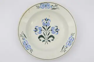 Farfurie cu fundul plat, puțin adâncit. Pe fundul vasului decor ilustrând un buchet de flori albastre cu tulpini și frunze verzi. Pe marginile vasului dispuse câte trei grupuri a două flori ca și cele de pe fund. La buză o linie neagră dispusă circular.. La posterior pe inelul de susținere practicate două perforații pentru atârnare. Cromatica: fond: alb; motive: albastru, verde, negru., 1850