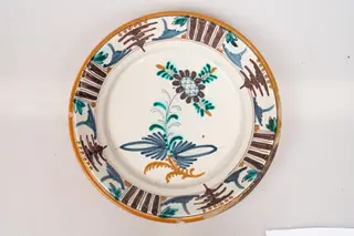 Farfurie de formă aplatizată, cu buza răsfrântă. Decorul, dispus pe un fond alb lăptos, prin pictare, este format dintr-un motiv floral stilizat, dispus central, pe fundul farfuriei, și motive geometrice (puncte galbene, grupuri de dungi paralele de culoare maro-brun, linii subțiri, paralele, linii sub formă de cerc, motivul bradului), dispuse într-un registru continuu pe buza farfuriei. Muchia buzei este marcată cu o dungă de culoare galbenă. Cromatica: alb, verde, brun, albastru, galben-ocru., 1850