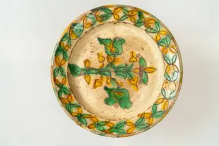 Farfurie de formă aplatizată, cu buză răsfrântă și muchie rotunjită. Piesa este modelată la roata olarului, angobată cu humă albă. Decorul este realizat prin sgrafitare în angoba crudă, peste care se aștern cu pensula culorile de smalț: verde și galben. Motivele decorative sunt florale și vegetale stilizate, dispuse pe fundul și pe buza farfuriei. Cromatica decorului: alb, galben, verde.