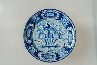 Farfurie de formă aplatizată, cu marginea lată, răsfrântă, ușor concavă spre interior. Decorul foarte bogat, dispus pe un fond alb, prin pictare, acoperă întreaga suprafață a farfuriei și este format din motive florale și vegetale fin stilizate (flori și frunze), avimorfe și un vas cu fructe (central, pe fundul farfuriei). Cromatica decorului: alb, albastru.
