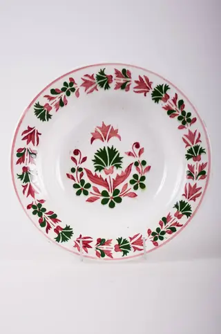 Farfurie de formă tronconică, adâncită; pe fund are găuri pentru agățare, Este decorată cu motive florale și geometrice; pe margine este decorat cu flori încadrate la exterior de o linie; în centru are mai multe flori. Pe fund are o marcă indescifrabilă. Cromatică: fond: alb; motive: vișiniu; verde., 1850