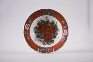Farfurie de formă tronconică din faianță, cu buza dreaptă, ornamentată prin pictare. Fundul este alb și are pictate trei flori roșii înconjurate de frunze mărunte, este delimitat de buza farfuriei cu o linie subțire, neagră. Buza vasului este roșie, decorată cu flori roz și frunzulițe discrete. În partea posterioară prezintă o gaură prin care se trece ața ce servește la agățarea farfuriei. Cromatică: fond: alb, roșu; motive: verde; roz; albastru; roșu; negru.