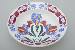 Farfurie de formă tronconică, ușor adâncită. Există două registre ornamentale, unul plasat de jur împrejurul buzei cu motive florale. Al doilea registru plasat în centru tot cu motive florale. Smălțuită. Cromatică: fond: alb; motive: albastru; verde; roșu; galben.
