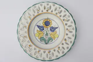 Farfurie ornamentală ce prezintă găurele între buză și partea adâncită obținute prin ștanțare. Buza este vălurită și prezintă un contur de culoare verde. Registrul central are motive florale și geometrice: cercuri galbene și albastre ce încadrează motivele florale. La posterior, baza inelară perforată pentru atârnare. Cromatică: fond: alb; motive: verde; galben; albastru; negru.