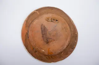 Farfurie plată cu baza largă. Marginea este decorată cu o linie vălurită albastră, iar în patru locuri cu câte un buchețel alcătuit dintr-un boboc verde cu frunze galbene dispus orizontal și intercalat de motivul bradului stilizat realizat prin hașurare. Decorul central reprezintă un buchet floral trasat cu cornul, alcătuit din două rodii mari maro, stilizate, o garoafă galbenă cu frunze și doi boboci galbeni de garoafă. Cromatică: alb, albastru, verde, galben, maro., 1850