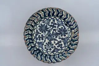 Farfurie semi-adâncă, cu fund larg, buză evazată și muchia buzei ondulată. Este acoperită pe suprafața interioară cu un strat de angobă albă peste care este pictat cu pensula și cu cornul decorul monocromatic format din motive geometrice și florale stilizate: registrul central de pe fundul farfuriei conține un buchet format din trei flori cu petale rotunde și mijlocul umplut cu linii hașurate intersectat, una dintre flori având mijlocul dintr-o linie spiralată, și din frunze; acest registru este încadrat între două linii circulare simple și o linie circulară ondulată și este completat cu șase motive formate din grupuri de linii crescătoare și descrescătoare, dispuse sub formă de triunghi și cu puncte. Registrul decorativ marginal de pe buza farfuriei este decorat cu un șir continuu de frunze duble între care se găsesc motive punctate (linii). Muchia buzei este marcată cu o dungă albastră. Suprafața ornamentată a farfuriei este acoperită cu un strat de smalț transparent. Cromatica: alb, albastru.