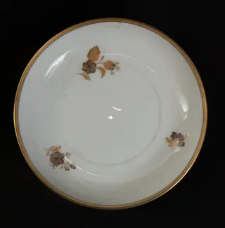 Farfurioară din porțelan alb, pictată manual cu un decor floral de culoare maro , ceașca este decorată pe margine cu auriu. Farfurioara face parte dintr-un serviciu de porțelan oferit cadou lui Nicolae Ceaușescu cu ocazia vizitei oficiale întreprinse în iunie 1973 în RFG.