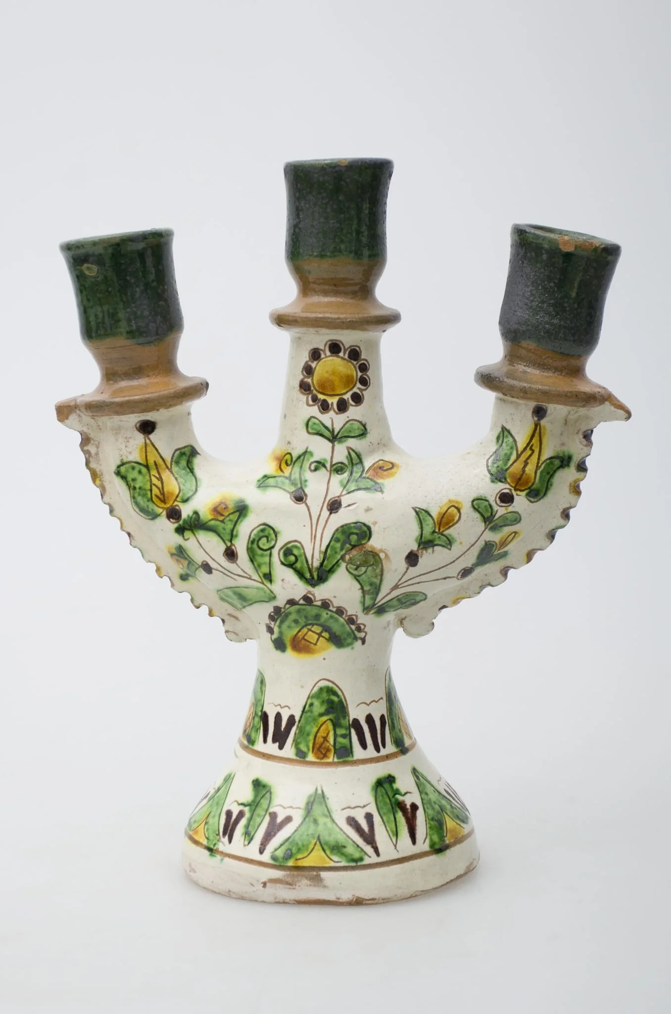 Obiect ceramic de uz casnic, format dintr-un picior tronconic, din care pornesc trei brațe, în care se introduc lumânările. Decorul pictural, realizat pe toată suprafața obiectului, este compus din motive geometrizante și vegetale. Primul registru, situat pe baza evazată, este format din elemente vegetale, dispuse circular și încadrat între două linii concentrice subțiri. Cel de-al doilea registru prezintă decor similar celui din registru inferior. Registrul superior este compus din trei flori, dintre care două (laterale) cu pistilul pronunțat, cea de-a treia, poziționată central, floarea soarelui. Gâtul și buza vasului au fost decorate cu verde și maro deschis. Iscălitură: indescifrabil. Cromatică: fond: alb; motive: crem; verde, maro închis, maro deschis, galben, negru., detail