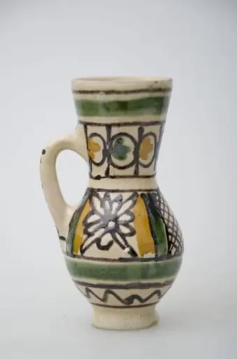 Vas ceramic de formă bitronconică, smălțuit de mici dimensiuni, cu baza plată și bombat în zona „pântecelui”. Prezintă o toartă ce pornește din zona gâtului redus ca dimensiuni și se termină în zona diametrul maxim al vasului. Decorul vasului este compus din două registre picturale. În primul registru, mărginit de două linii drepte subțiri, de culaore maro închis, au fost pictate două flori conturate cu negru și încadrate de câte două linii groase de culoare verde și galben. Cel de-al doilea registru este compus din boboci de culoare verde și galben, fiind dispuși concentric pe gâtul vasului. Ambele registre picturale sunt încadrate între două linii groase circulare, verzi. Pe toartă a fost redactat textul: „KORUND”. Prezintă uzură funcțională. Cromatică: fond: alb; motive: galben, verde, maro închis., by Iosza Ianos