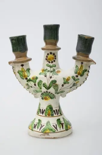 Obiect ceramic de uz casnic, format dintr-un picior tronconic, din care pornesc trei brațe, în care se introduc lumânările. Decorul pictural, realizat pe toată suprafața obiectului, este compus din motive geometrizante și vegetale. Primul registru, situat pe baza evazată, este format din elemente vegetale, dispuse circular și încadrat între două linii concentrice subțiri. Cel de-al doilea registru prezintă decor similar celui din registru inferior. Registrul superior este compus din trei flori, dintre care două (laterale) cu pistilul pronunțat, cea de-a treia, poziționată central, floarea soarelui. Gâtul și buza vasului au fost decorate cu verde și maro deschis. Iscălitură: indescifrabil. Cromatică: fond: alb; motive: crem; verde, maro închis, maro deschis, galben, negru., 1850