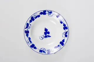 Obiect de uz casnic de formă tronconică. Pe fond alb se regăsește un ornament albastru: central două frunze de viță de vie. Pe margine farfuria este decorată cu frunze de viță de vie cu vrej și cu un cerc albastru. Cromatică: fond: alb; motive: albastru.