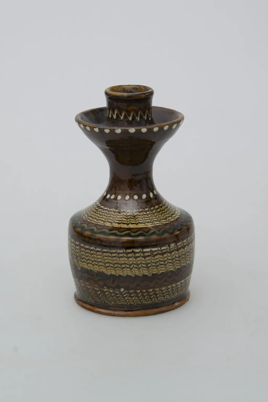 Recipient ceramic tubular, folosit ca suport de lumânare. Baza, fără fund, foarte înaltă cu pereții drepți ce se continuă printr-un gât îngust, înalt, lățindu-se în partea superioară pe marginea care se evazează la exterior. Este prevăzut un șanț pentru ținerea cerii topite. Partea superioară prevăzută cu un orificiu înalt pentru fixat lumânarea. Decorații jirăvite pe întreg corpul, linie verde ondulată pe umăr, linie punctiformă circulară la baza gâtului și pe inelul reliefat, și o linie în zig-zag în partea superioară a gâtului. Smălțuit în exterior. Cromatică: fond: maro închis; motive: alb; verde; maro; negru; bej., by Ogrezeanu Stelian