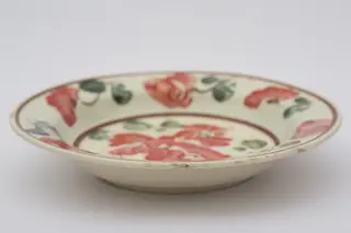 Piesă ceramică smălțuiță de dimensiuni reduse și formă cilindrică, cu baza plată și susținută de un inel circular, pereții ușor ridicați și buza larg evazată. Suprafața interioară a recipientului a fost ornamentată cu două registre decororative cu motive geometrizante și fitomorfe stilizate. Central, pe fundul vasului au fost pictați trei flori roșii cu frunze verzi, cel mai probabil maci. Marginal, pe bordura farfuriei a fost realizat cel de-al doilea registru pictural, format din șase maci cu frunze verzi, dispuși circular. Cel de-al doilea registru este încadrat de două linii circulare drepte, de culoare mov. Farfuria era folosită în gospodărie pentru decorarea interioarelor. Prezintă uzură funcțională. Cromatică: fond: alb; motive: roșu; verde; mov.