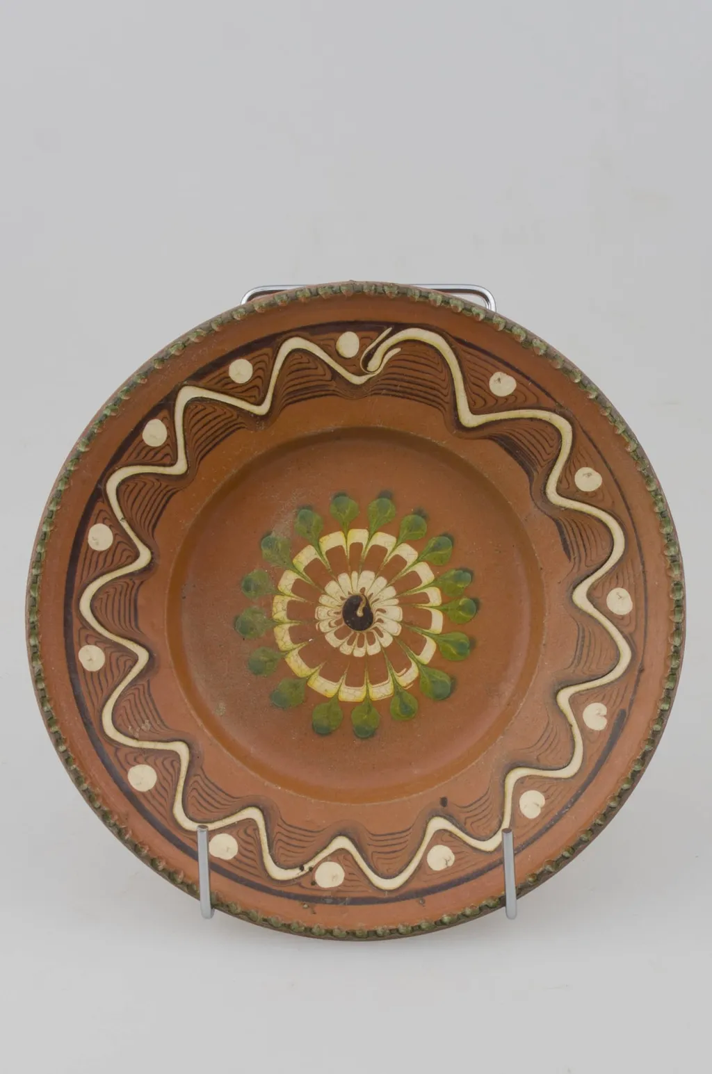 Farfurie cu pereții rotunjiți, marginea oblică lată și buza crestată. Prezintă decor geometric pe margine: linii frânte și curbe; pete de culoare; central avem o floare stilizată. Cromatică: fond: maro; motive: verde; alb., by Răducanu Ion