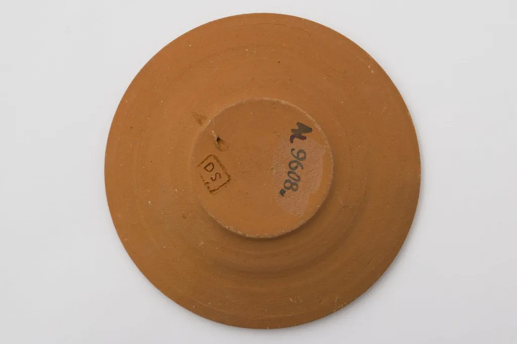 Vas ceramic cu fundul plat puțin adâncit și marginile evazate, puțin ridicate. Ornamentată cu pensula cu angoba albă. Fundul este marcat printr-o spirală din care ies alte șase spirale mici dispuse pe circumferința exterioară. Pe marginea vasului două benzi, una cu o linie întreruptă, a doua cu șase valuri întrerupte. La posterior ștanțat ”DS” încadrat într-un dreptunghi. Cromatică: fond: cărămiziu; motive: alb., by Șchiopu Dumitru