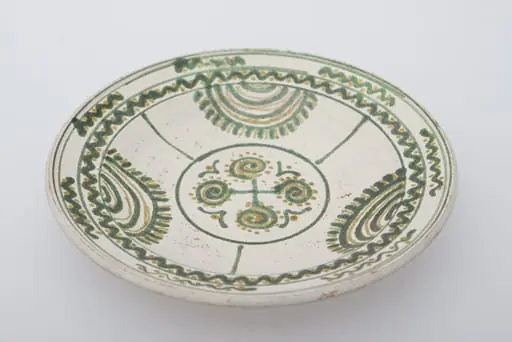 Vas ceramic de formă circulară și profil tronconic. Fața farfuriei a fost introdusă în angobă albă. Decorația a fost făcută cu pensula. Pe fundul farfuriei un cerc circumscris patru motive în spirală. Pereții farfuriei, au fost împărțiți în trei părți egale, pe circumferință, delimitate fiecare parte de o linie verticală, motivul bradului este reprezentat printr-o succesiune de semicercuri, pe ultima dintre ele pictându-se "țepii" bradului. Buza farfuriei a fost împărțită în două registre, în primul fiind pictat valul continuu, a doua cu val întrerupt. Pe fundul vasului, la spate, ștanțat ”D. Șchiopu C. Vlădești 1980'' înscris într-un cerc. Pe inelul de susținere dispuse două orifficii pentru atârnare. Cromatică: fond de angobă albă; motive: smalț verde și maro., by Șchiopu Dumitru