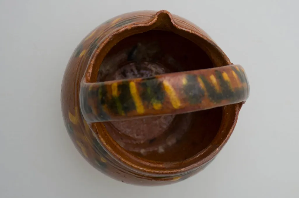 Vas de mici dimensiuni sub forma de bijou; pântecos cu gâtul scurt și gros, gură răsfrântă și cioc. Smălțuit în interior și la exterior. În partea superioară se află două linii incizate. Pe corp avem motive florale stilizate, iar pe mănușă decor geometrice: liniar și punctiform. Diametral, peste gură aven o mânușă aproximativ la fel de înaltă ca vasul. Cromatică: fond: cărămiziu; motive: galben, verde., by Șchiopu Dumitru