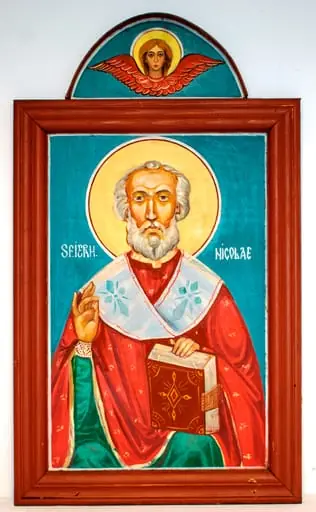Sfântul ierarh Nicolae este redat 3/4, așezat. Binecuvântând cu mâna dreaptă, cu stânga sprijinind o Evanghelie pe genunchi. Pictat cu capul decoperit, fruntea înaltă, figura sobră, expresivă, bine proporționată. Părul și barba albe, scurte, ondulate. Ochii căprui, nasul drept.Înveșmântat cu sacos roșu decorat cu motive florale miniaturale, stihar verde cu mânecuțe brodate cu perle, epitrahil alb cu cruci gri. În jurul capului aureolă de culoare galbenă, cu chenar dublu(roșu și alb). Evanghelia este legată în piele, cu coperta decorată cu motive geometrice pe fond cafeniu. Fond bleu închis, cu o linie de chenar albă, subțire, la fel cu inscripția scrisă în dreptul umerilor. Paleta cromatică este variată, dar nu foarte armonioasă, culorile reci. Pictura este realizată de un pictor contemporan cu școală, autorul picturii unor biserici și icoane. Rama profilată, lată, alcătuită din patru stinghii îmbinate oblic la colțuri, bătută în cuie metalice, culoare brun roșcat. În partea de sus este adăugată o porțiune semicirculară, cu margine din placaj îngust, montat transversal. Pe fondul bleu închis al acesteia este pictat un heruvim cu aripile întinse, chip sobru, cu păr blond, ochi negri gura roșie. În partea de jos sunt două cuie mari de fier cu care era fixată.