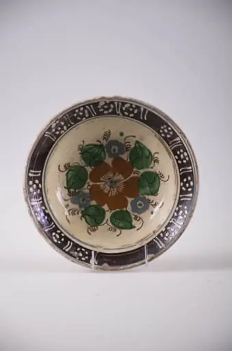 Blid de formă tronconică cu partea superioara usor rasfrântă, Decorul este pictat cu pensula cu un motiv central de tip floral; pe margini linii paralele și flori stilizate din puncte. Smălțuit. Cromatică: roșu; verde și maro în centru; motive laterale: alb pe fond brun inchis; fond general: alb-galbui., by Unknown