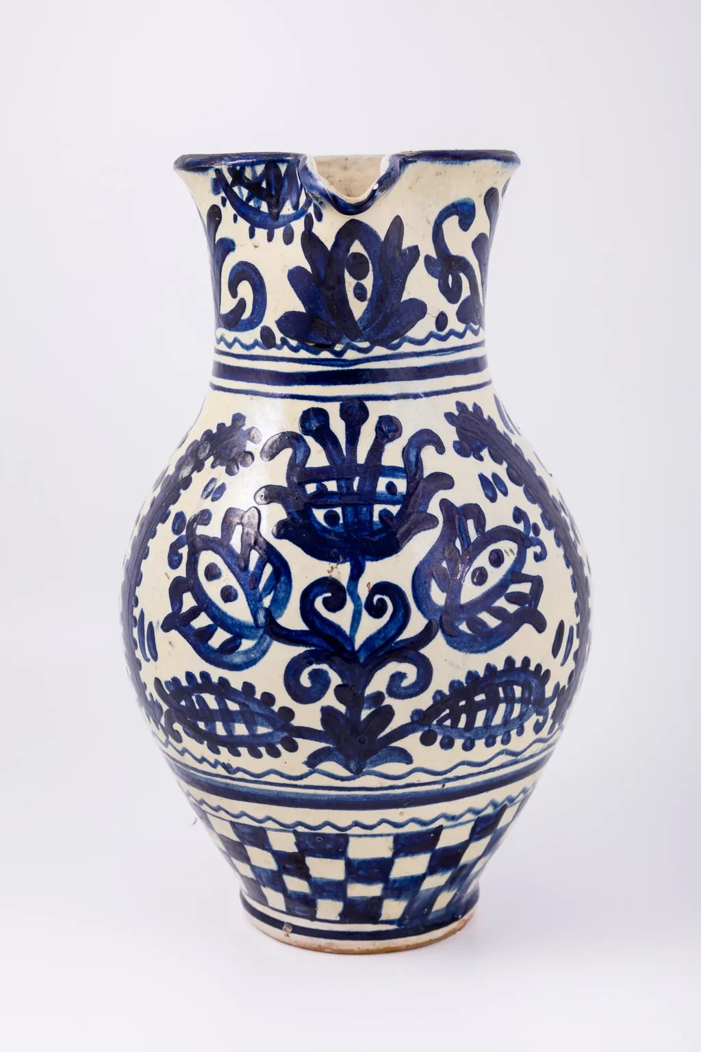 Cană piriformă prevăzută cu o toartă dreaptă și lată, gura rotundă, prevăzută cu cioc de scurgere. Suprafața decorativă a vasului este împărțită în patru registre decorative. Primul registru dispus pe gâtul piesei prezintă un decor floral și fitomorf reprezentând lalele și lujeri. Al doilea registru prezintă o linie vălurită și trei linii subțiri circulare. Decorul central, reprezintă trei „arbori ai vieții” stilizați realizați din trei lalele mari, înflorite, lujeri și frunze, separați de câte o semilună. Registrul al patrulea prezintă motivul tablei de șah. Deasupra toarte pictate cu motive florale și fitomorfe este redat anul confecționării piesei: 1980, iar pe fundul vasului numele persoanei care a pictat piesa: Herta Wilk. Cromatica: alb, albastru-cobalt., by Unknown, unspecified, 1980