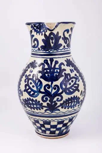 Cană piriformă prevăzută cu o toartă dreaptă și lată, gura rotundă, prevăzută cu cioc de scurgere. Suprafața decorativă a vasului este împărțită în patru registre decorative. Primul registru dispus pe gâtul piesei prezintă un decor floral și fitomorf reprezentând lalele și lujeri. Al doilea registru prezintă o linie vălurită și trei linii subțiri circulare. Decorul central, reprezintă trei „arbori ai vieții” stilizați realizați din trei lalele mari, înflorite, lujeri și frunze, separați de câte o semilună. Registrul al patrulea prezintă motivul tablei de șah. Deasupra toarte pictate cu motive florale și fitomorfe este redat anul confecționării piesei: 1980, iar pe fundul vasului numele persoanei care a pictat piesa: Herta Wilk. Cromatica: alb, albastru-cobalt., by Unknown
