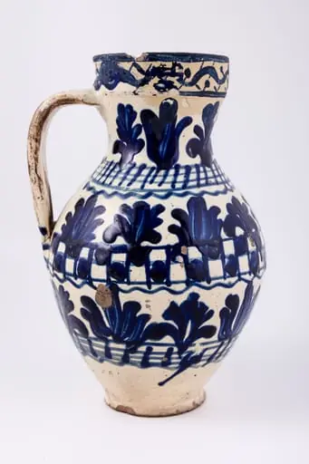Cană piriformă prevăzută cu o toartă dreaptă și lată, gura rotundă. Suprafața decorativă a vasului este împărțită în șapte registre decorative: cel din partea superioară, pe buza vasului este dispus orizontal, iar celelalte șase, dispuse vertical. Primul registru, așezat pe marginea dreaptă a cănii redă o linie vălurită intercalată de puncte. Registrul al doilea, al patrulea și al șaselea prezintă motive fitomorfe și florale, brăduți intercalați de lalele înflorite stilizate. Registrul al treilea, dispus pe gâtul cănii și al șaptelea, dispus la bază, prezintă linii circulare orizontale intersectate de linii verticale. Registrul al patrulea prezintă tabla de șah. Toarta este pictată cu pensula. Cromatica: alb, albastru-cobalt., by Unknown