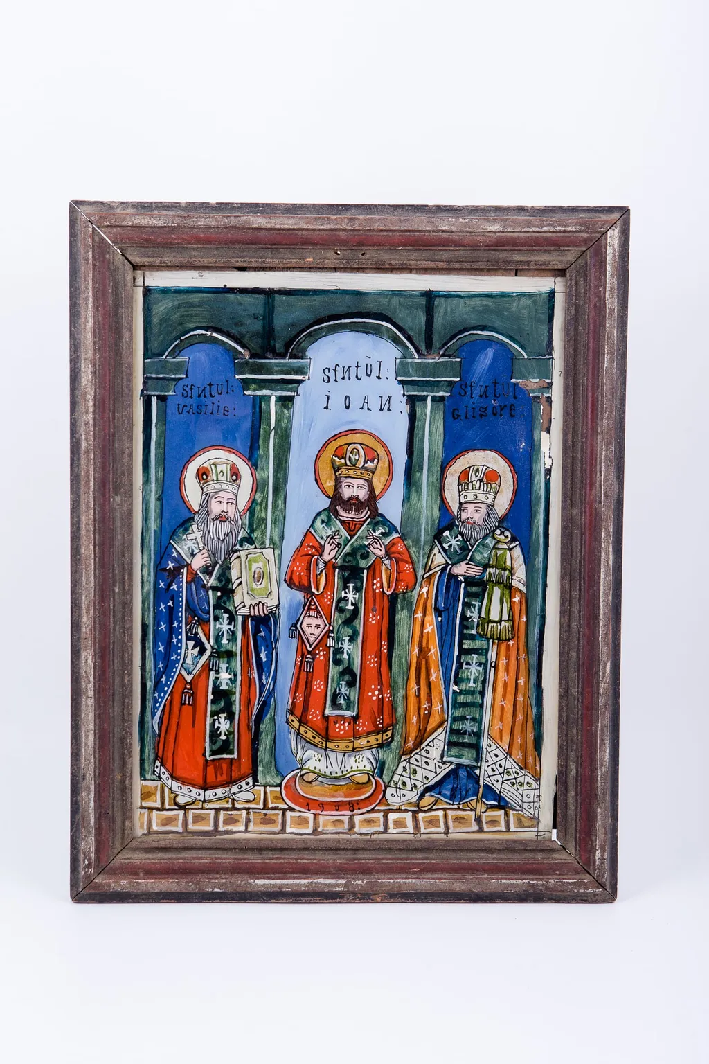 Cei Sfinți Trei Ierarhi Vasile cel Mare, Grigorie de Dumnezeu Cuvântătorul și Ioan Gură-de-Aur slujesc și săvârșesc Sfânta Liturghie împreună. Sunt înveșmântați în veșminte liturgice: sacos, felon și epitrahil mithră și aureolă. Cromatica veșmintelor este vie și luminoasă: roșu vermillon, orange-cărămiziu, galben-ocru și albastru ceruleum și bleu-ciel, verde și alb. Sfântul Vasile cel Mare are crucea în mâna dreaptă și binecuvântează și în mâna stângă ține Sfânta Evanghelie închisă. Sfântul Ioan Gură-de-Aur, central, tânăr cu barba brunetă, binecuvântează cu ambele mâini și are pe bedernița romboidală un chip de sfânt, element antrpomorf. Alături este Sfântul Grigorie care ține în mâna stângă cârja-sceptrul arhiepiscopală și binecuvântează. Fondul întregii tematici iconografice este albastru ceruleum în exterior și central bleu-ciel. Sfinții trei ierarhi sunt încadrați de coloane și arcade-bolți, schițându-se astfel structura arhitecturală de biserică. În registrul inferior este un un caroiaj alb-ocru, o lespede de fundație puternică - reprezentând credința creștin-ortodoxă. Cromatica este luminoasă și armonioasă: roșu vermillon, orange, verde spanac-avocado, galben-ocru, brun, albastru ceruleum și bleu-ciel, gri, alb și negru. Este o icoană de Valea Sebeșului. Inscripiția din româno-chirilică se traduce: Sfântul Vasilie Sfântul Ioan Sfântul Grigorie. Icoana este datată 1908., by Unknown, 1908