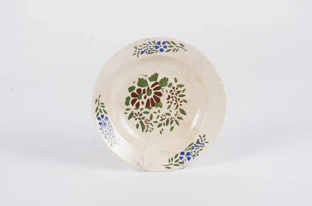 Farfuria este de formă tronconică, fiind smălțuită integral. Prezintă un decor floral-vegetal dispus pe fundul și pe buza vasului. Cromatică: fond alb; motive: verde; maro; albastru., by Unknown, 1850